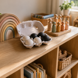 A wooden toy shelf in a nursery or playroom setting, featuring a wooden rainbow arch toy, a cardboard egg carton with black and white eggs, and wooden peg dolls on a tray.