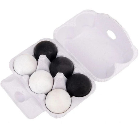 A white styrofoam egg carton holding six eggs, four black and two white, photographed on a plain white background.