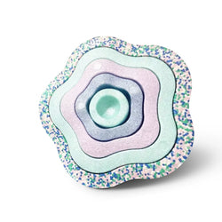 A flower-shaped baby pacifier clip made of layered textured materials, featuring concentric rings in pastel mint green, lavender, light blue, and a glittery multicolor outer edge, shown on a white background.